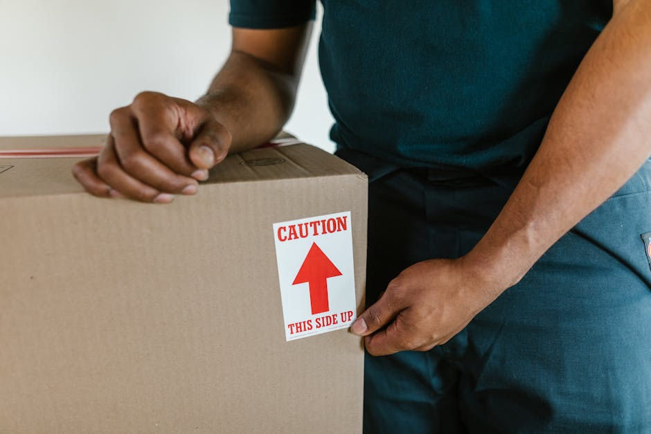 Close-up of a person wearing a dark green shirt and navy trousers, holding the edge of a large cardboard box during a home relocation. The box is sealed with packing tape and has a red and white caution label indicating 'Caution' with an upward arrow and the instruction 'This Side Up,' suggesting careful handling. The person’s hands are gripping the box firmly, positioned on either side, as part of the packing and moving process. In the background, a plain wall and part of a door frame are visible, with the box situated on a flat surface, likely inside a property or a loading area. The lighting is neutral, emphasizing the professional nature of the packaging activities carried out by Movers Acton, a company specialising in removals and furniture transport in the Acton area, aligned with services such as house moves near Acton Town station.
