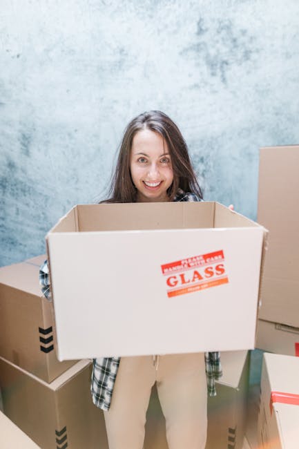 Two women engaged in a home relocation, standing amidst numerous packed cardboard boxes of various sizes, some sealed with red tape and others open with packing materials visible. One woman, wearing a checkered shirt and beige trousers, holds a medium-sized box, while the other, dressed in a white t-shirt and patterned pants, carries a large ceramic vase with a leafy green plant. They are inside a room with a textured grey wall and wooden flooring, surrounded by the boxes positioned near a doorway or open area for loading. The scene captures the process of packing and organizing items for a move, with the women smiling and preparing to load the boxes onto a vehicle such as a van, in line with professional house removal services provided by Movers Acton for local relocations, including aspects of furniture transport and packing logistics.