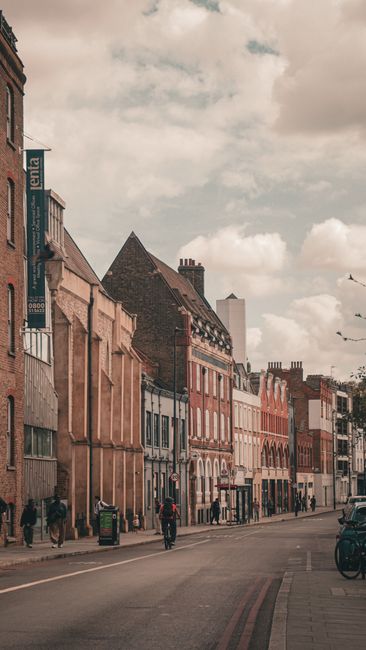 A street view in Acton with a row of older terraced houses featuring brick facades, some with decorative window frames and pitched roofs. The image captures a partly cloudy sky overhead, with a few pe
