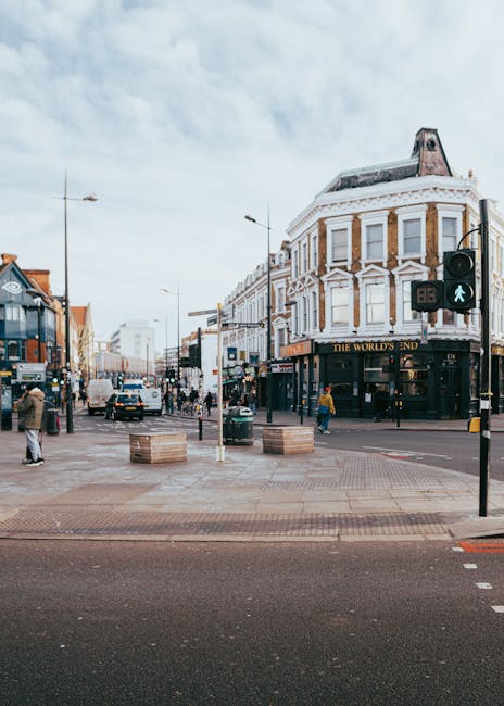 A street view in Acton with a row of older terraced houses featuring brick facades, some with decorative window frames and pitched roofs. The image captures a partly cloudy sky overhead, with a few pe